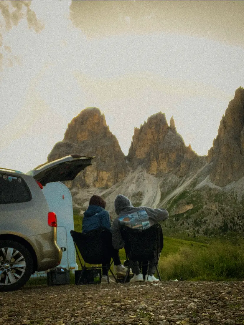 Two people sit on camping chairs next to the car, facing the rock towers of the Dolomites.
