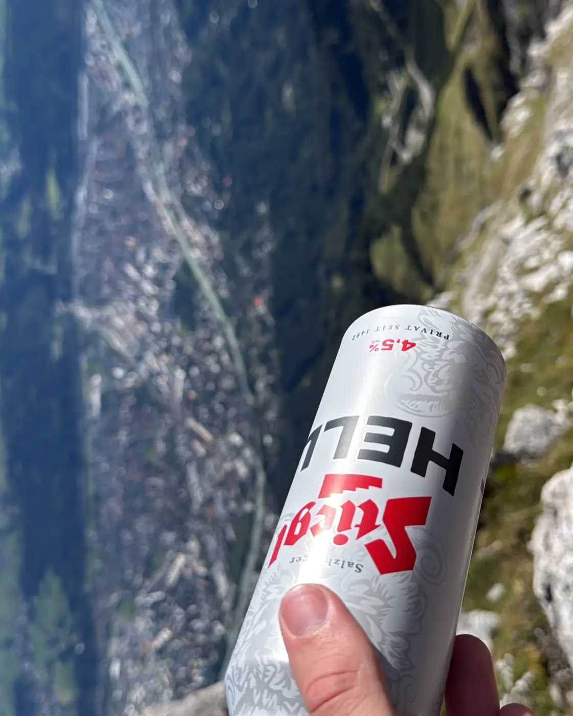 A hand holds up a can of Stiegl high above Innsbruck, with the valley far below.