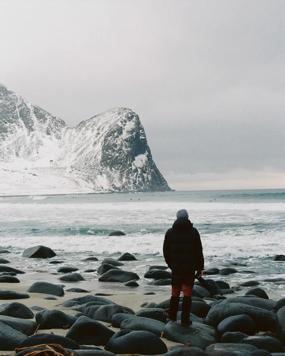 Someone stands on a stony beach in Norway, facing the sea and a snow-covered mountain.
