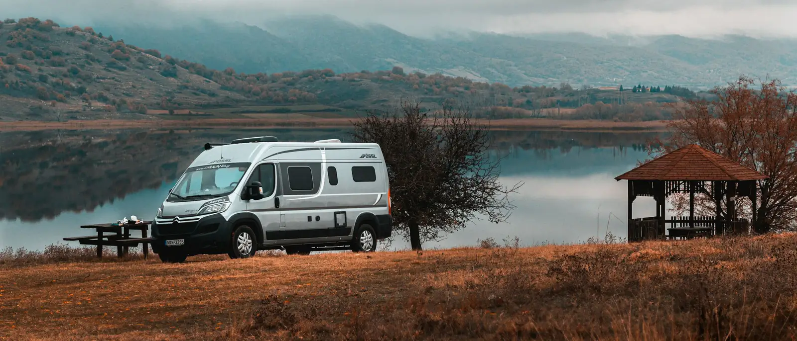 Ein Campervan steht am Morgen an einem See, davor eine Person mit einer Tasse