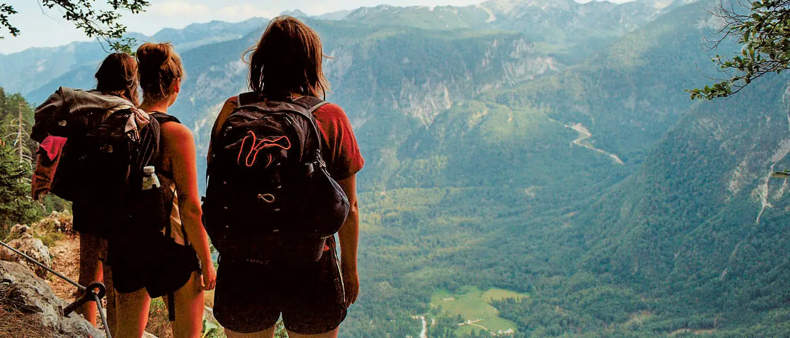 Three friends with backpacks stand on a mountain path, seen from behind, looking into a wide valley