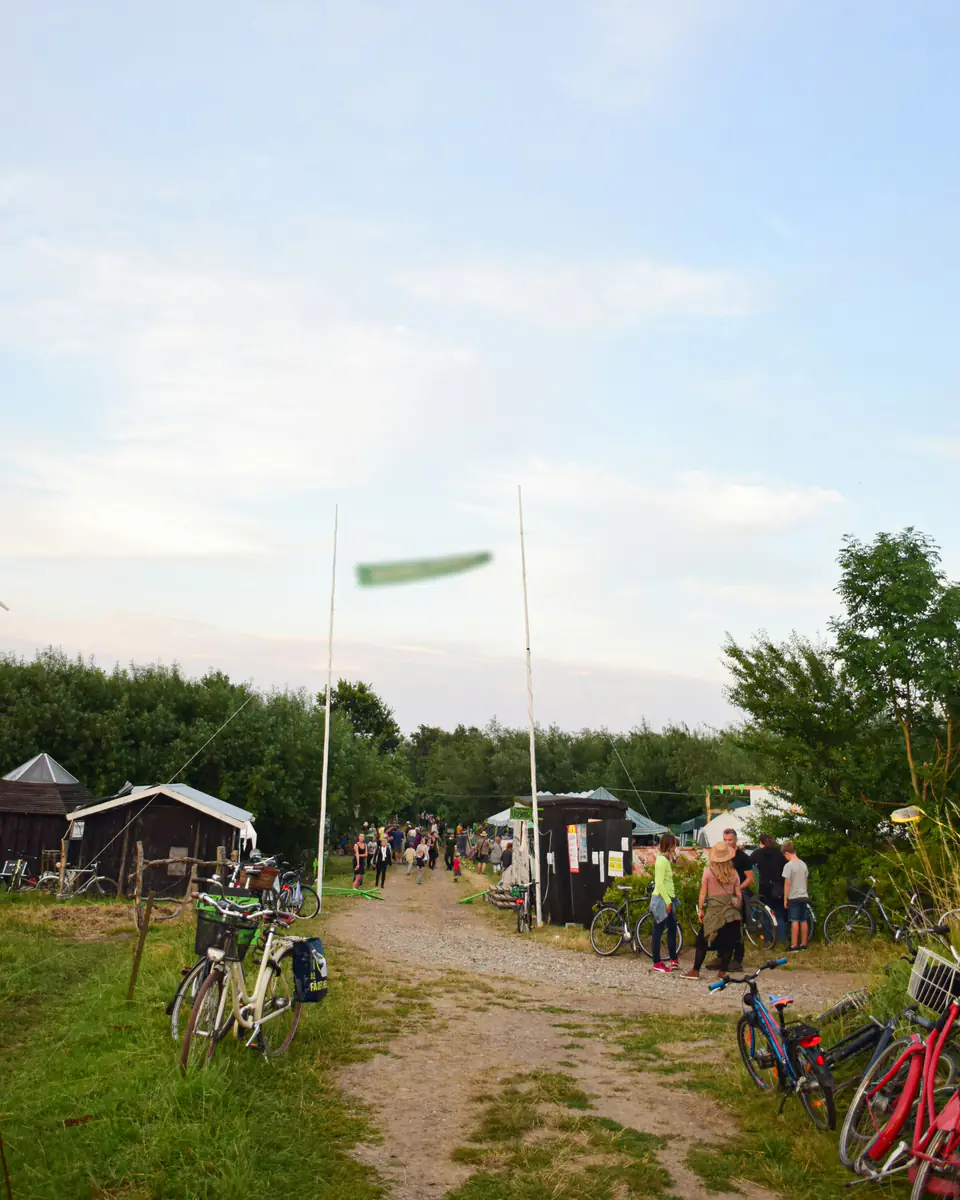 Eingang eines kleinen Festivals auf einer Wiese: ein Kiesweg zwischen zwei Fahnenmasten, Fahrräder am Rand, Gäste gehen hinein