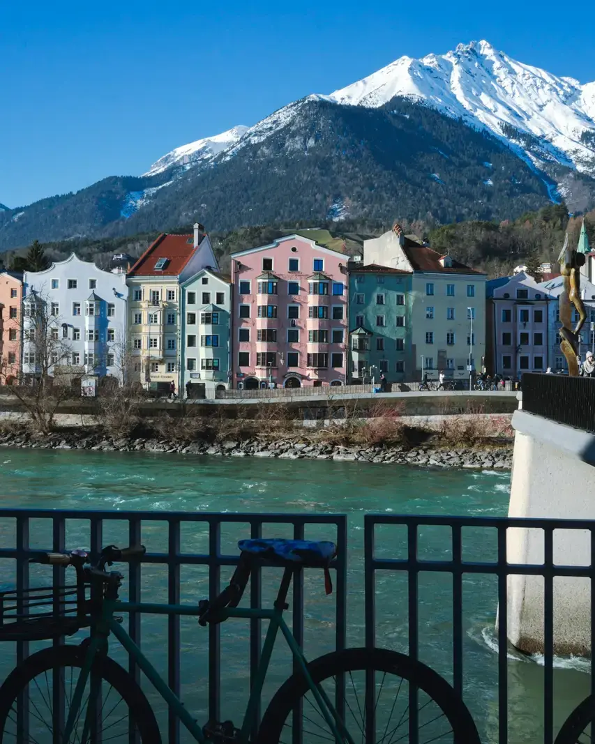 Ein Rad am Geländer vor bunten Häusern am Fluss, dahinter Berge
