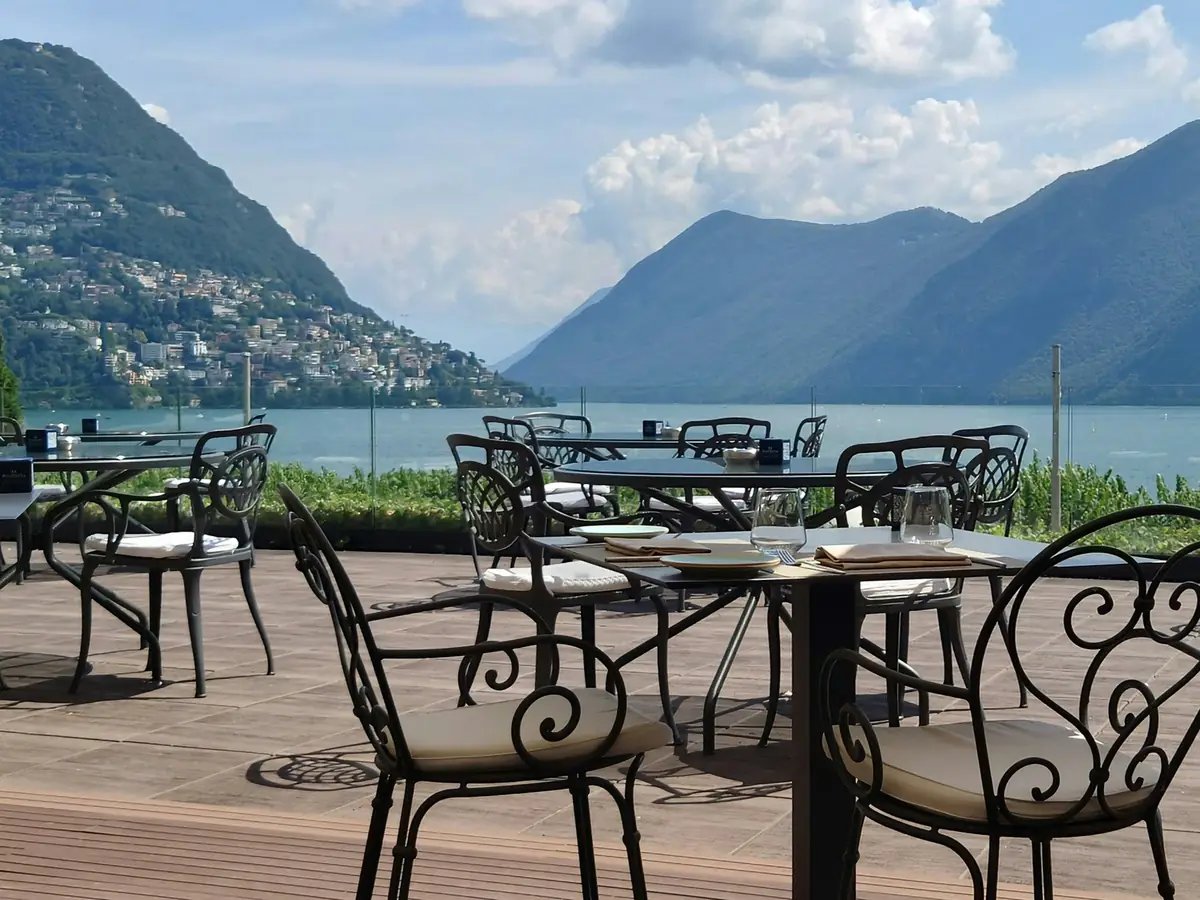 Restaurantterrasse über einem See mit gedeckten Tischen, dahinter Berge und ein Dorf am Hang