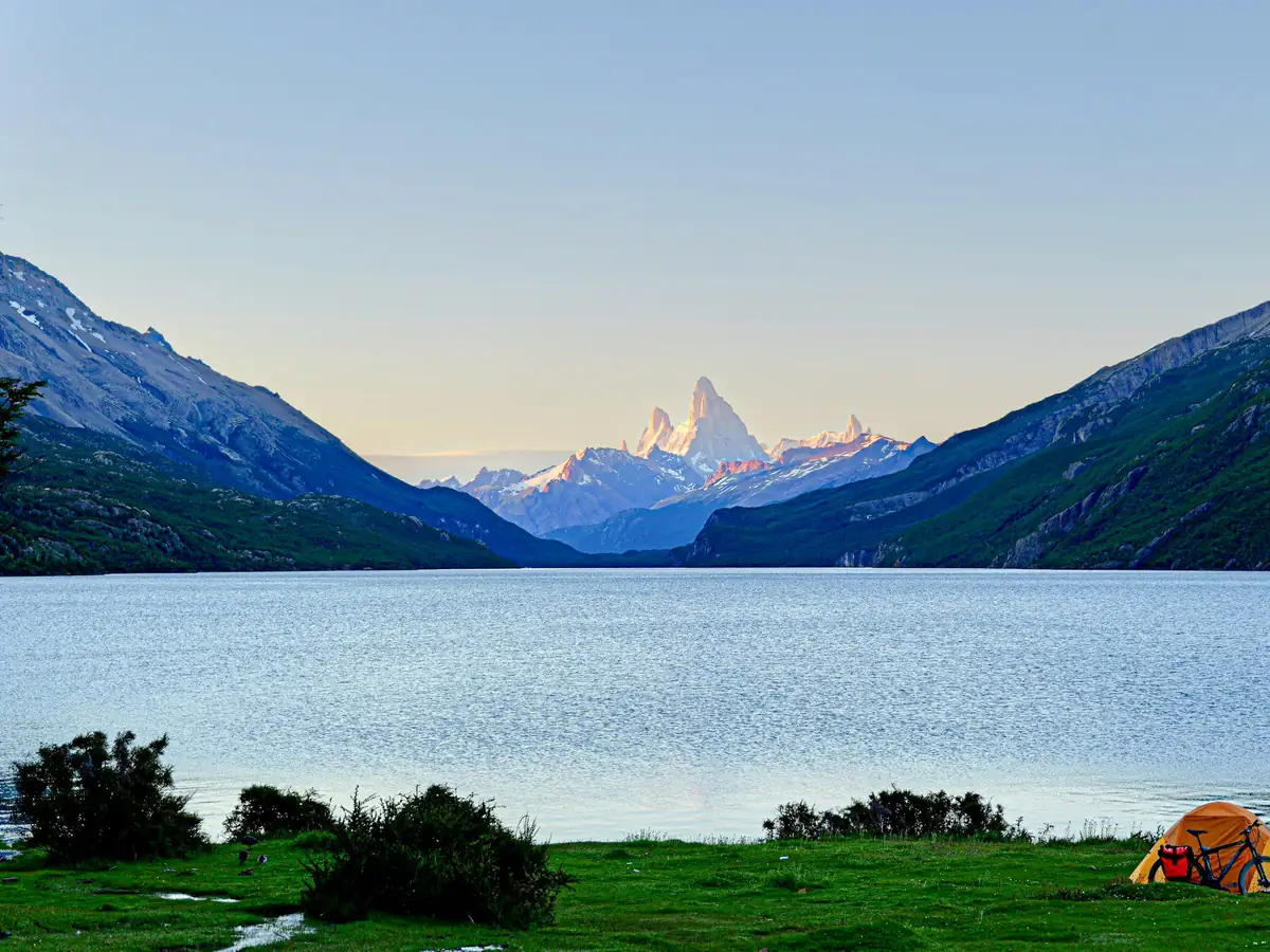 Ein Zelt am Ufer eines Bergsees, dahinter ein spitzer Gipfel im Morgenlicht