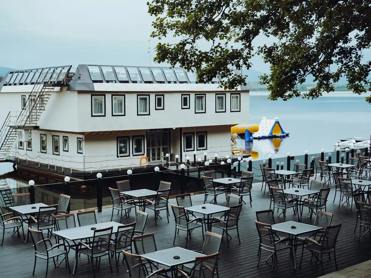 Leere Terrasse eines Cafés am See in der Morgensonne, dahinter Berge