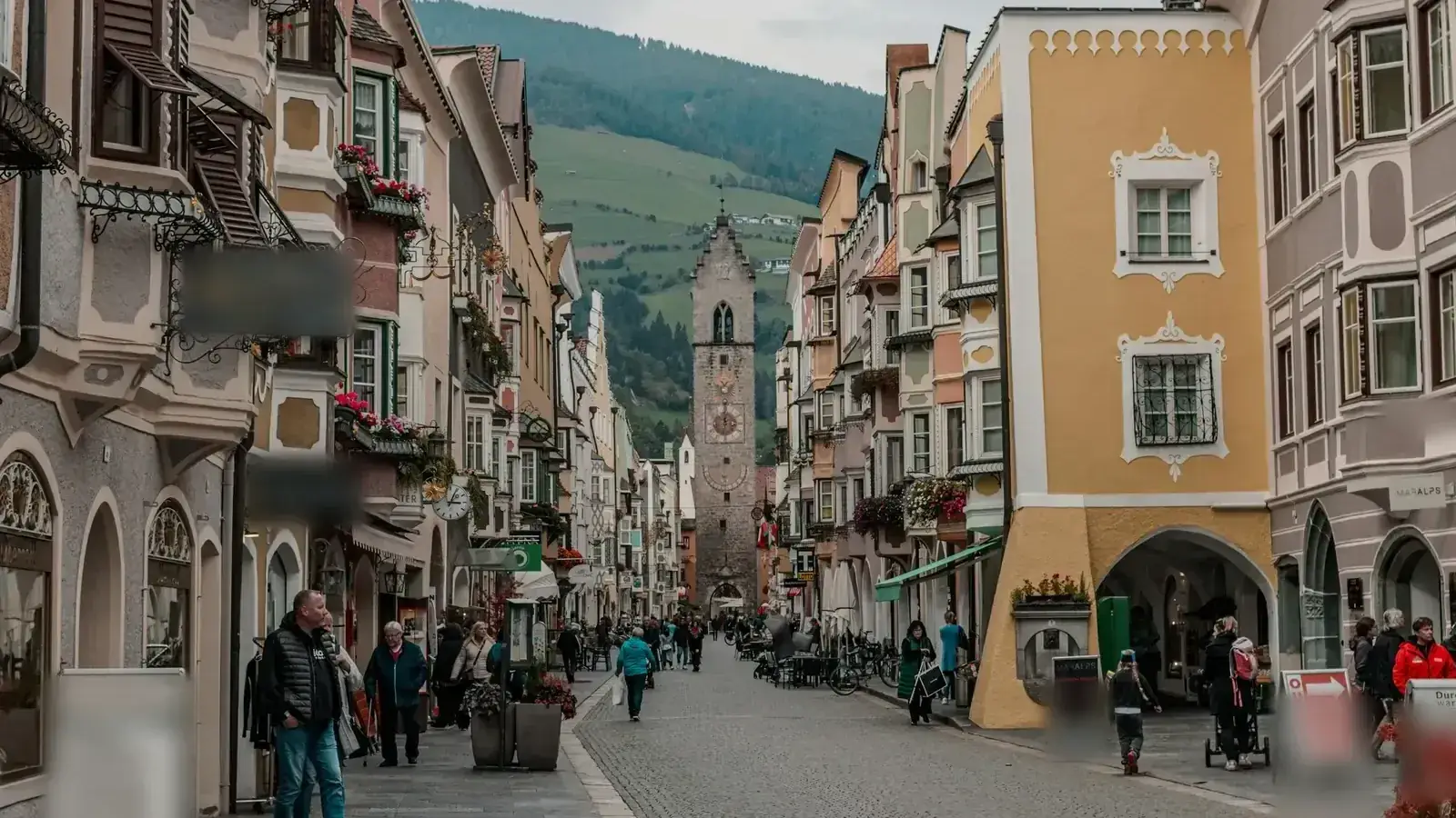 Altstadtgasse mit Läden bei Tag, Menschen klein im Bild, Berge im Hintergrund