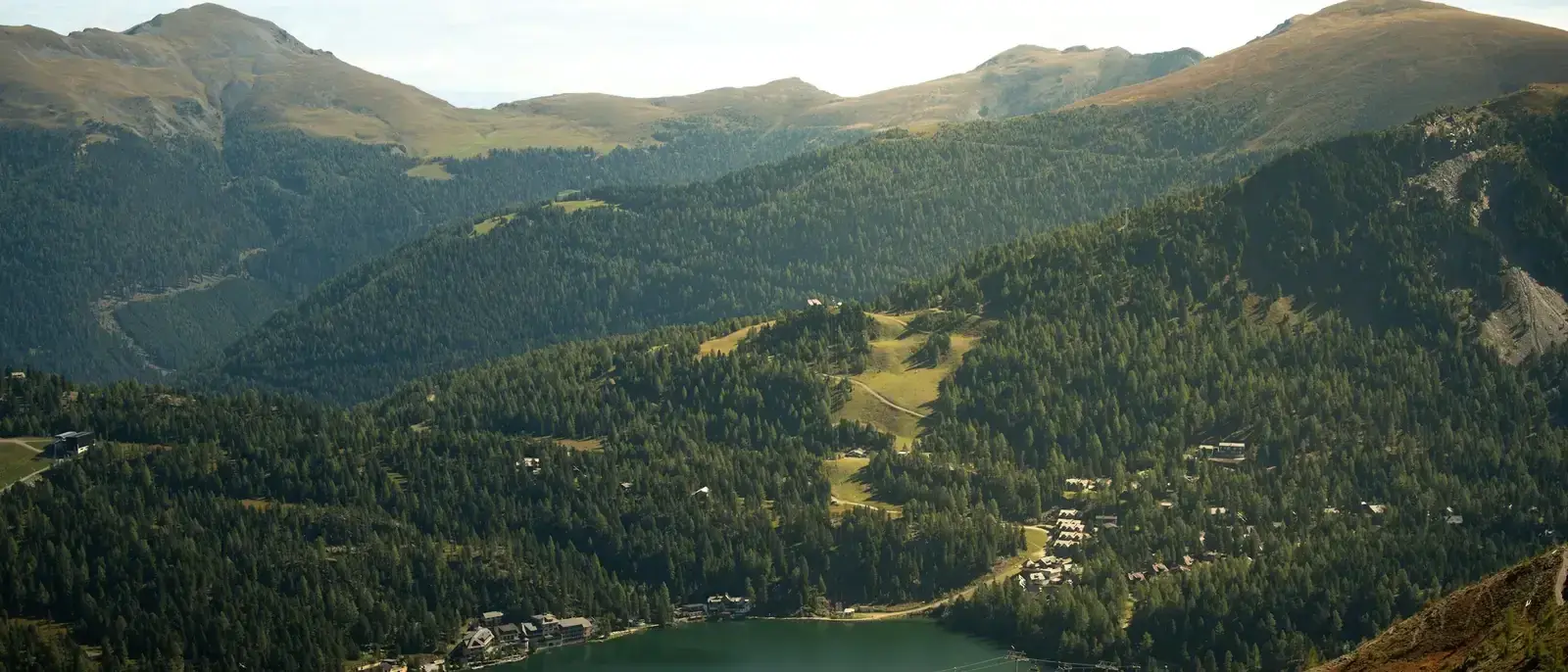Ein stiller Bergsee im Tal, umgeben von Wald und wenigen Häusern.