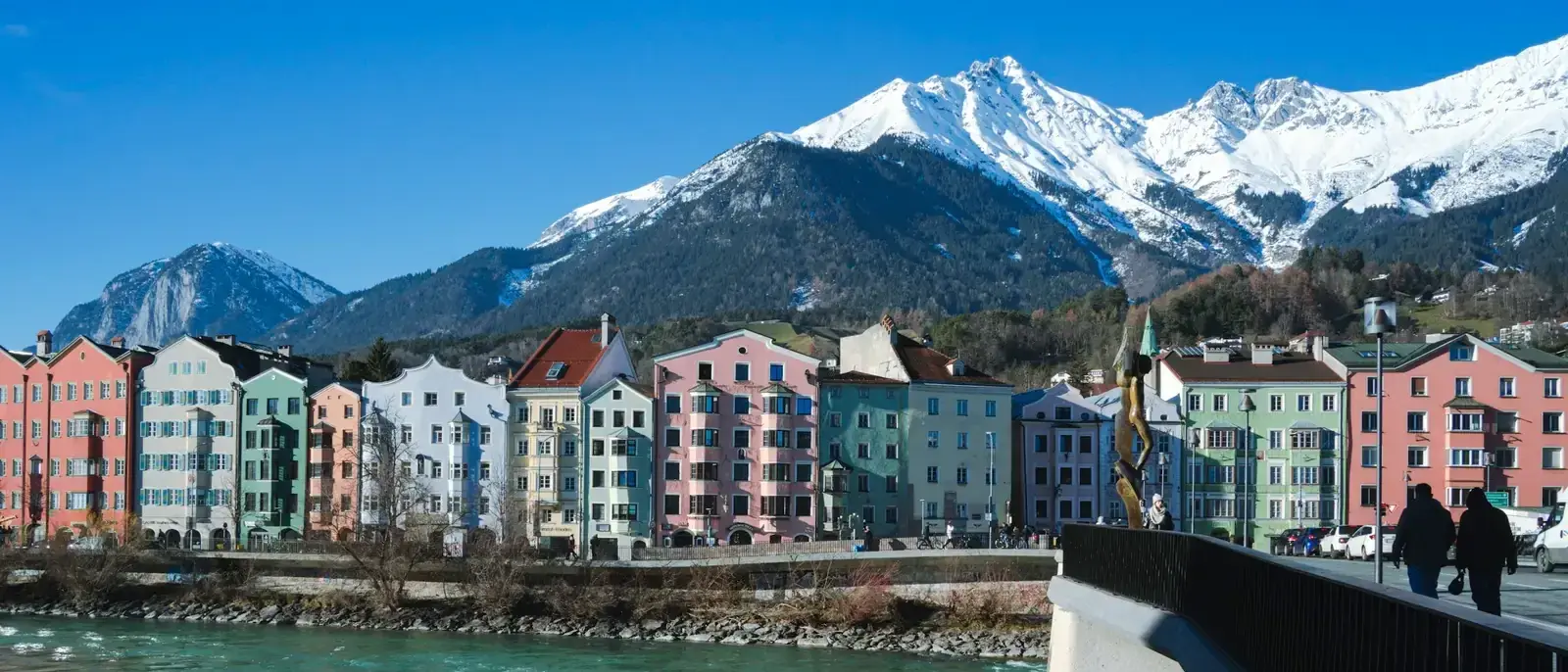 Bunte Häuser am Fluss vor schneebedeckten Bergen, im Vordergrund Fahrräder am Brückengeländer.